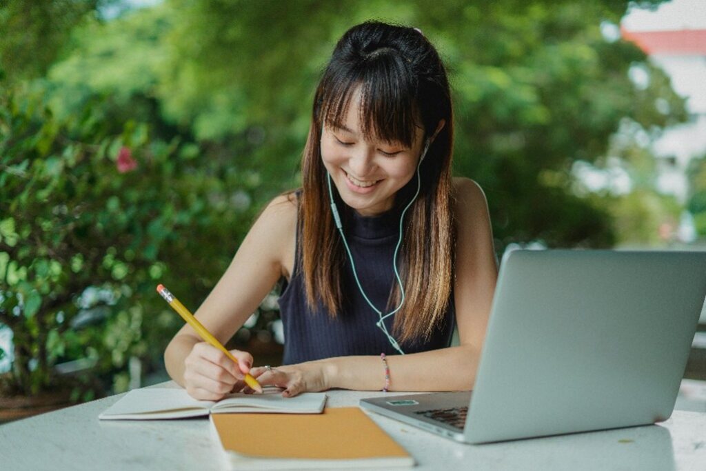 Jeune femme souriante assise en train de prendre des cours en ligne avec un ordinateur et un carnet de notes.